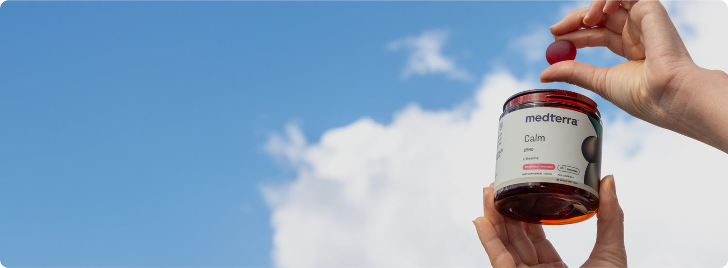 Hands holding a jar of medterra Calm 25MG L-theanine supplement with a red gummy above the jar against blue sky