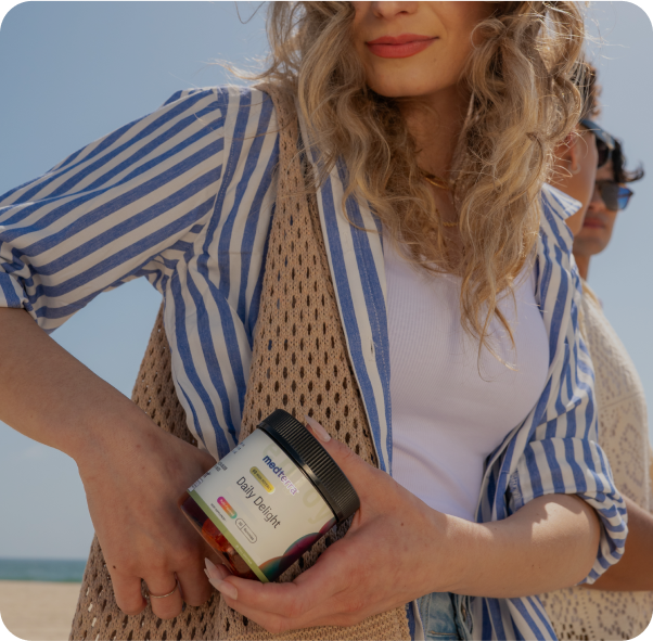 Person wearing blue-striped shirt and crochet bag on a beach, holding a jar labeled 'medterra Daily Delight'.