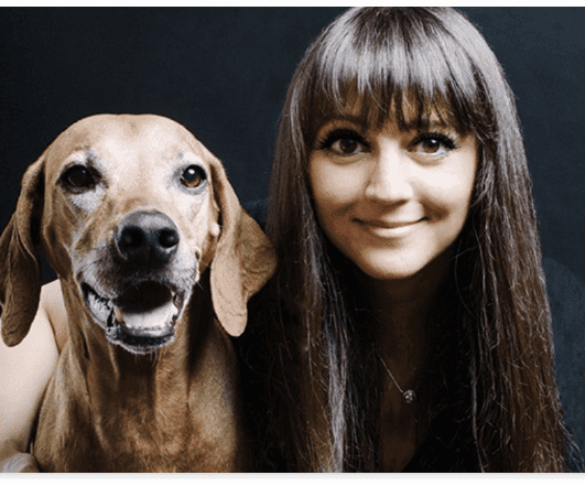 Smiling woman with long brown hair beside a seated brown dog, both looking toward the camera.