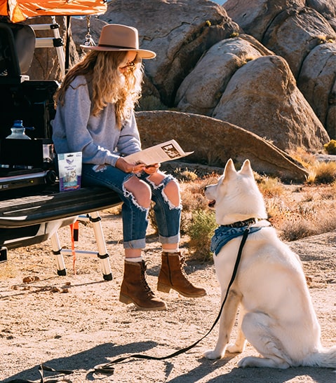 Woman seated at a vehicle tailgate in a rocky desert, reading a photo to a white dog wearing a blue bandana.