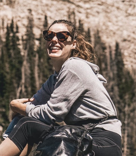 Smiling woman in sunglasses and gray hoodie sitting outdoors with trees and a rocky cliff behind her.